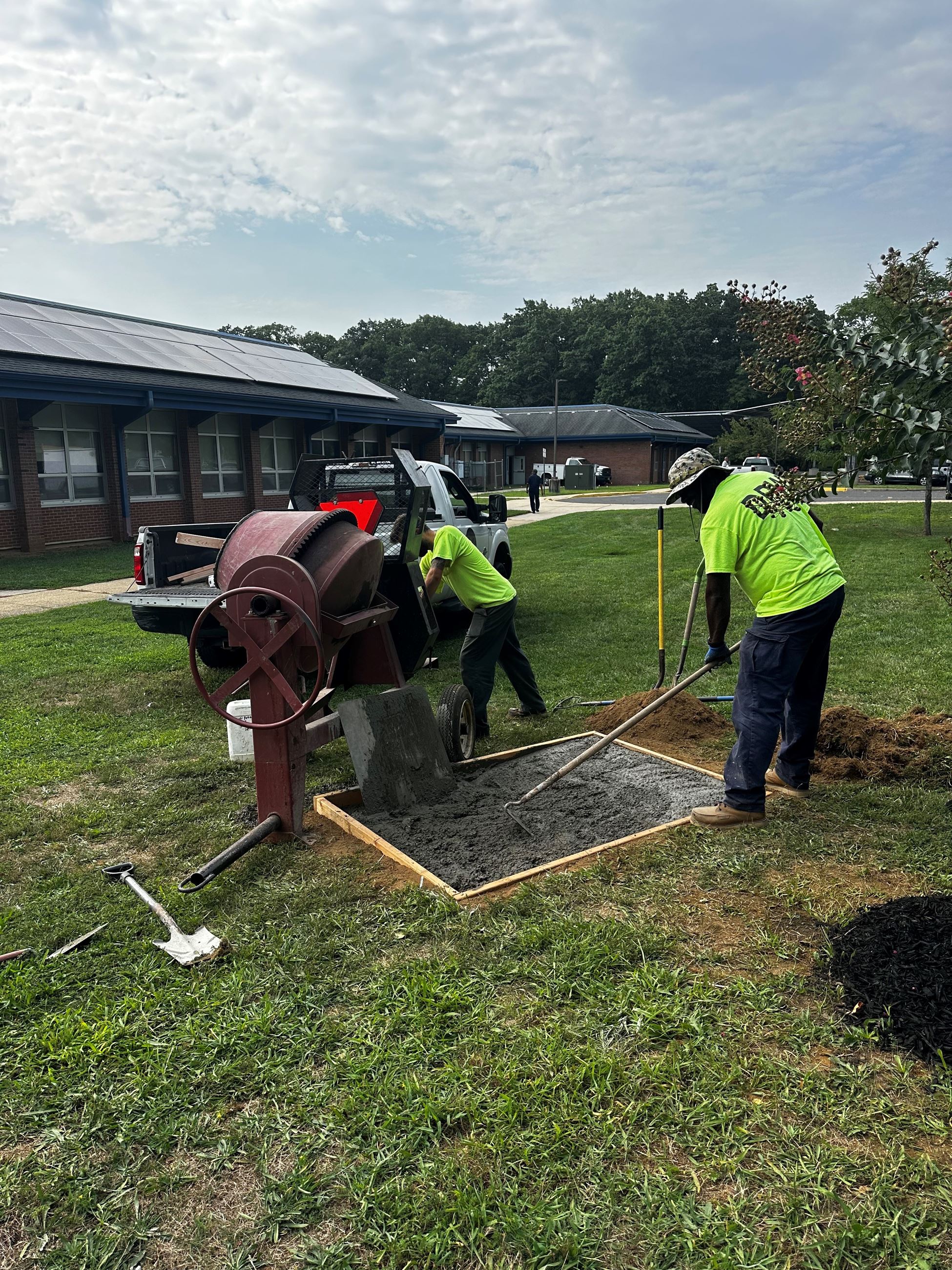Two workers in neon shirts operate a concrete mixer and shovel dark concrete into a bordered area outside brick buildings.